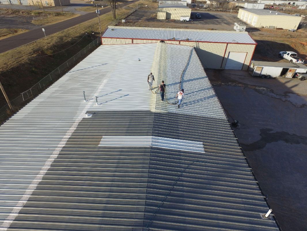 Workers painting a metal roof; one side is silver, the other is dark gray, on a sunny day.