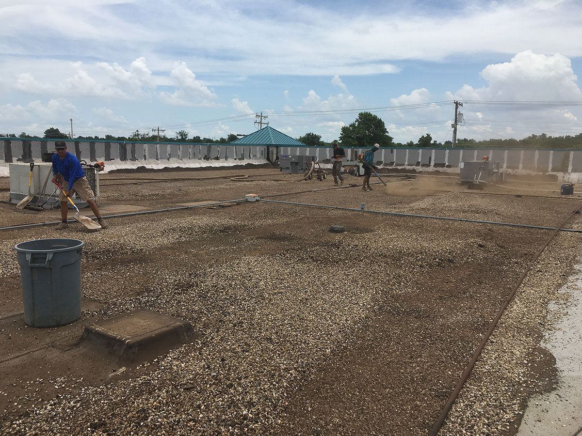 Workers spreading gravel on a rooftop under a cloudy sky. A blue trash can is in the foreground.