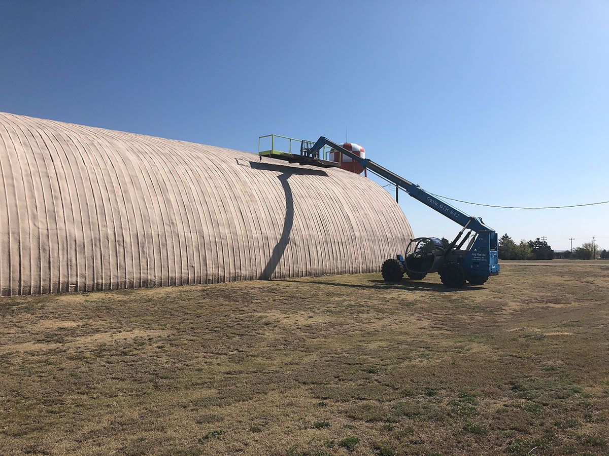 A worker on a lift repairs the roof of a large, arched, white building. Sunny, outdoor setting.