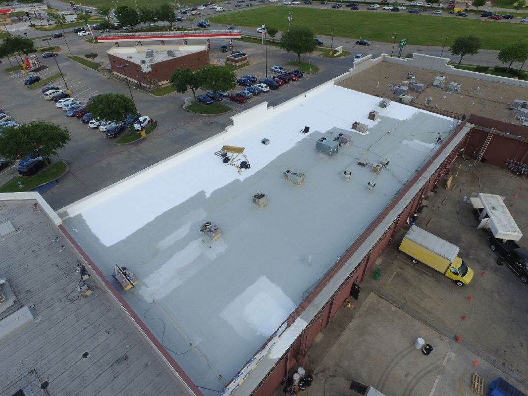 Aerial view of a commercial roof, partially coated in reflective white paint, with parking lot and buildings in the background.