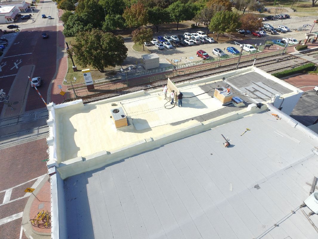 Aerial view of a flat roof with workers, newly applied foam, and nearby train tracks.