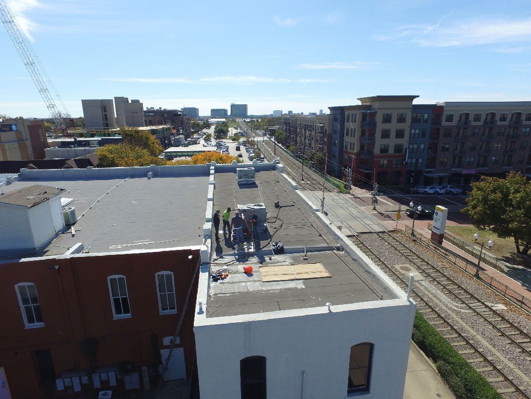 Aerial view of a flat rooftop with workers, city street, train tracks, and buildings on a sunny day.