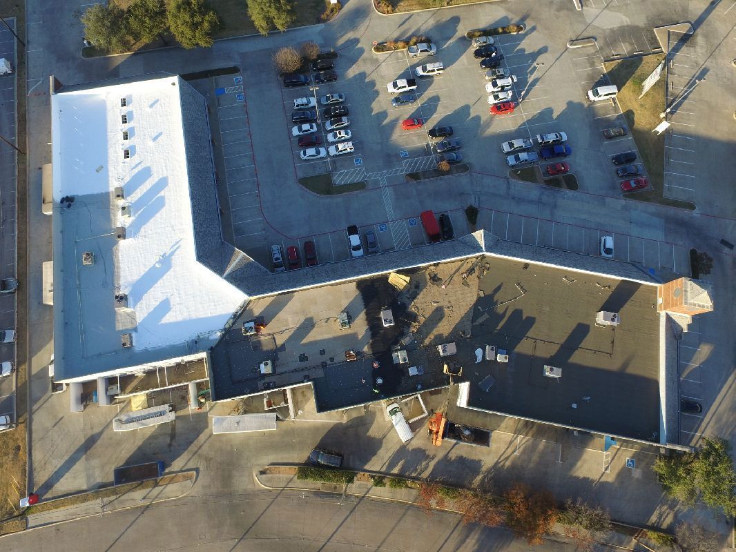 Aerial view of a building with a bright white roof, parking lot with cars, and trees in an outdoor setting.