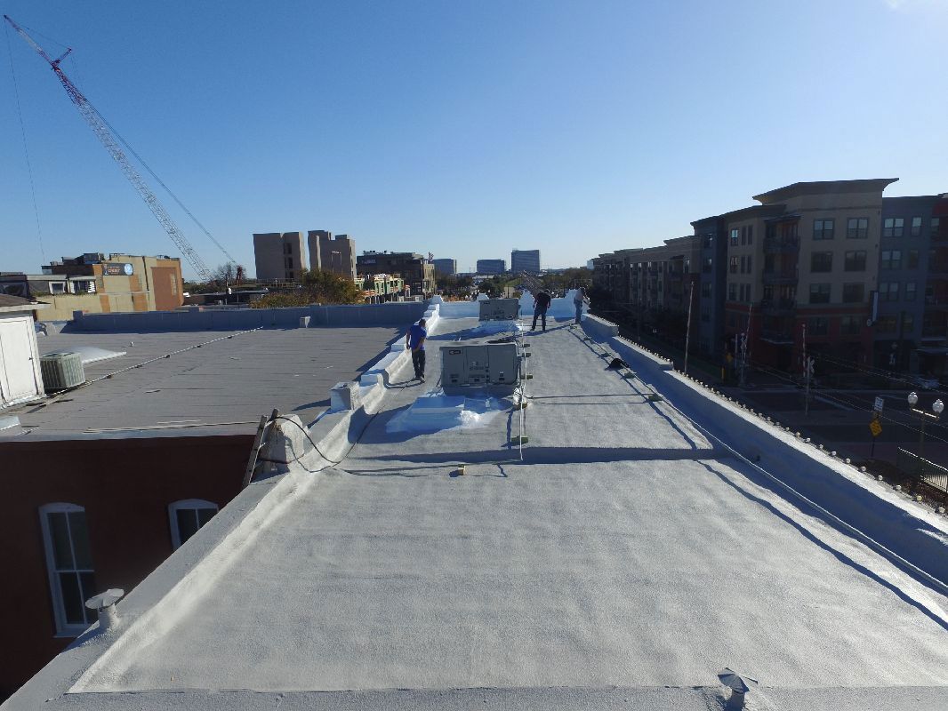 Rooftop view of flat gray roofing with workers. City buildings and blue sky in background.