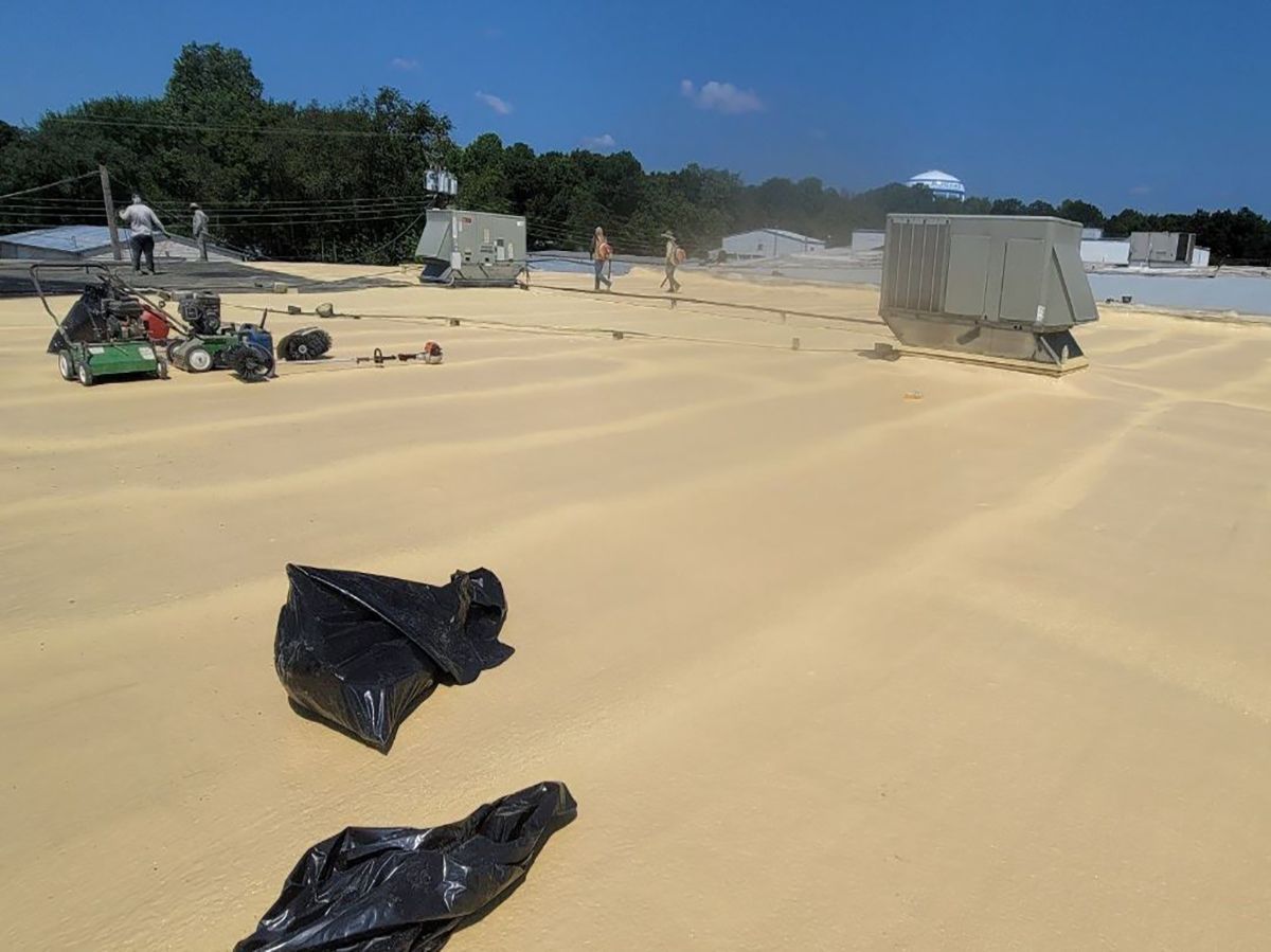 Workers apply yellow sealant to a flat roof on a sunny day, with equipment and trash bags visible.