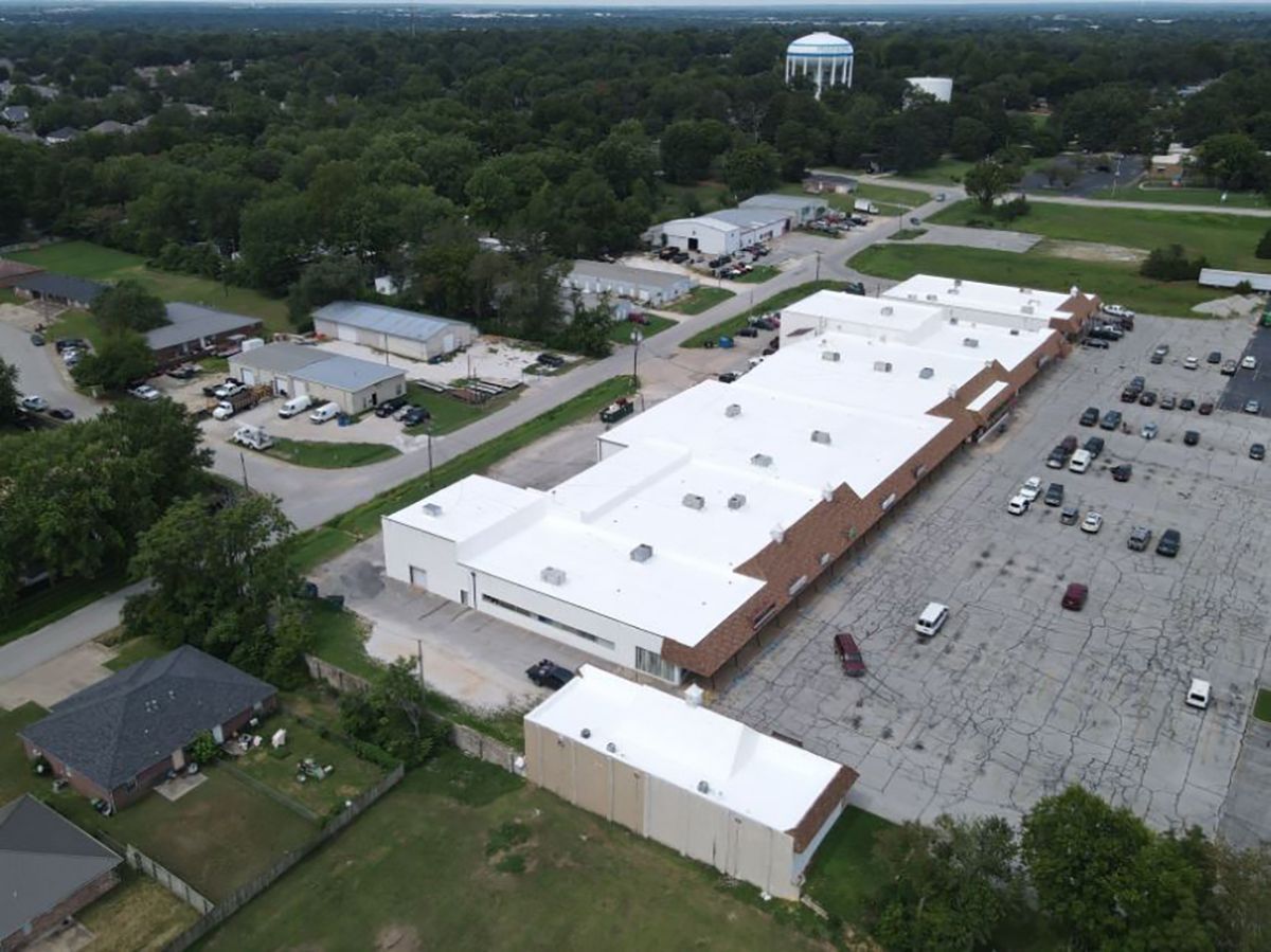 Aerial view of a large, white-roofed commercial building with a parking lot and surrounding green trees and buildings.
