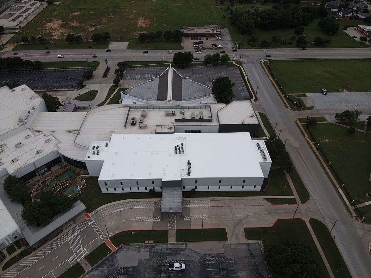 Aerial view of a large white building with a flat roof, surrounded by roads, parking lots, and green trees.