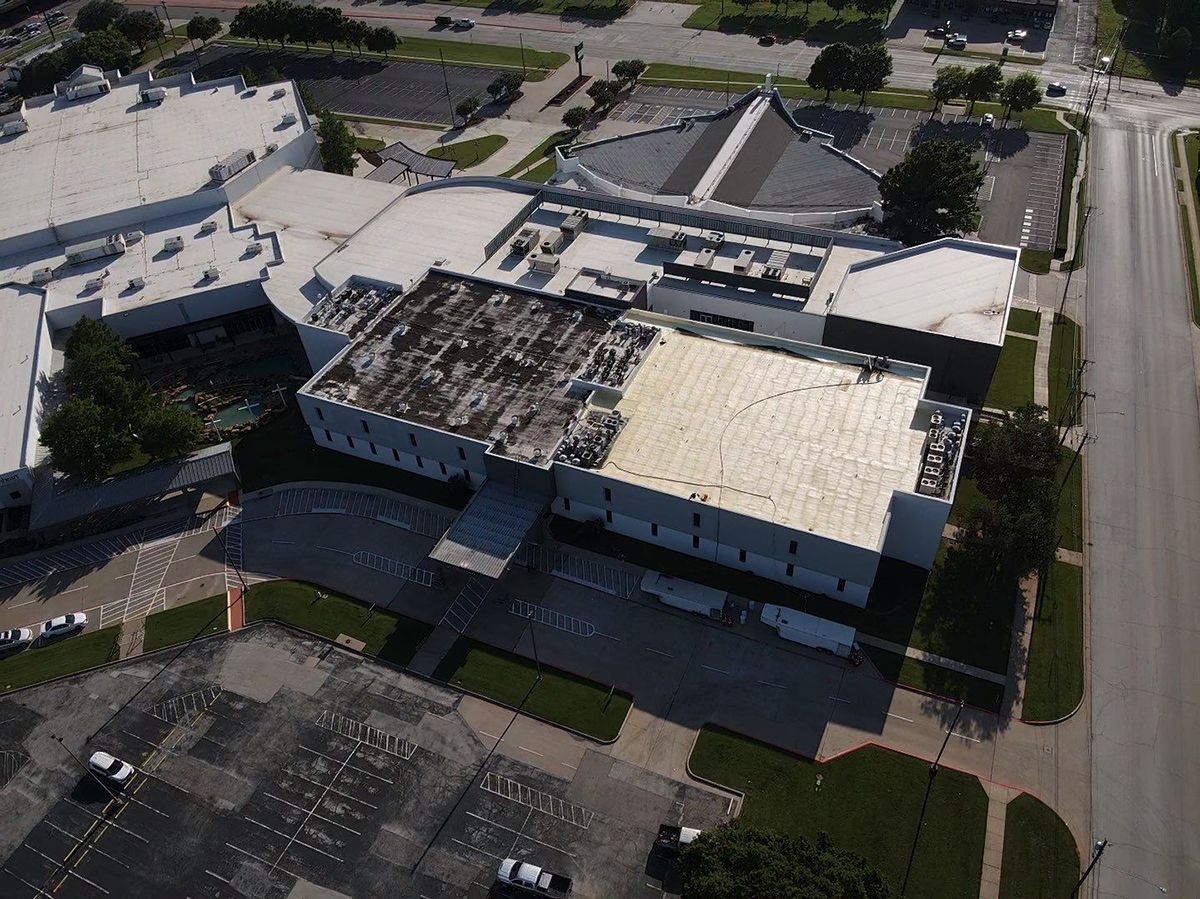 Aerial view of a large, light-colored building complex with a parking lot and street alongside it.