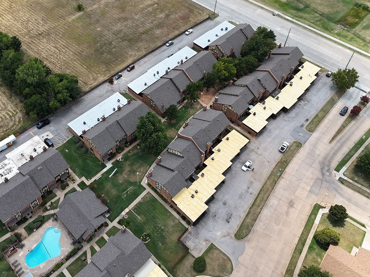 Aerial view of apartment buildings with brown roofs, carports, a swimming pool, and a road.