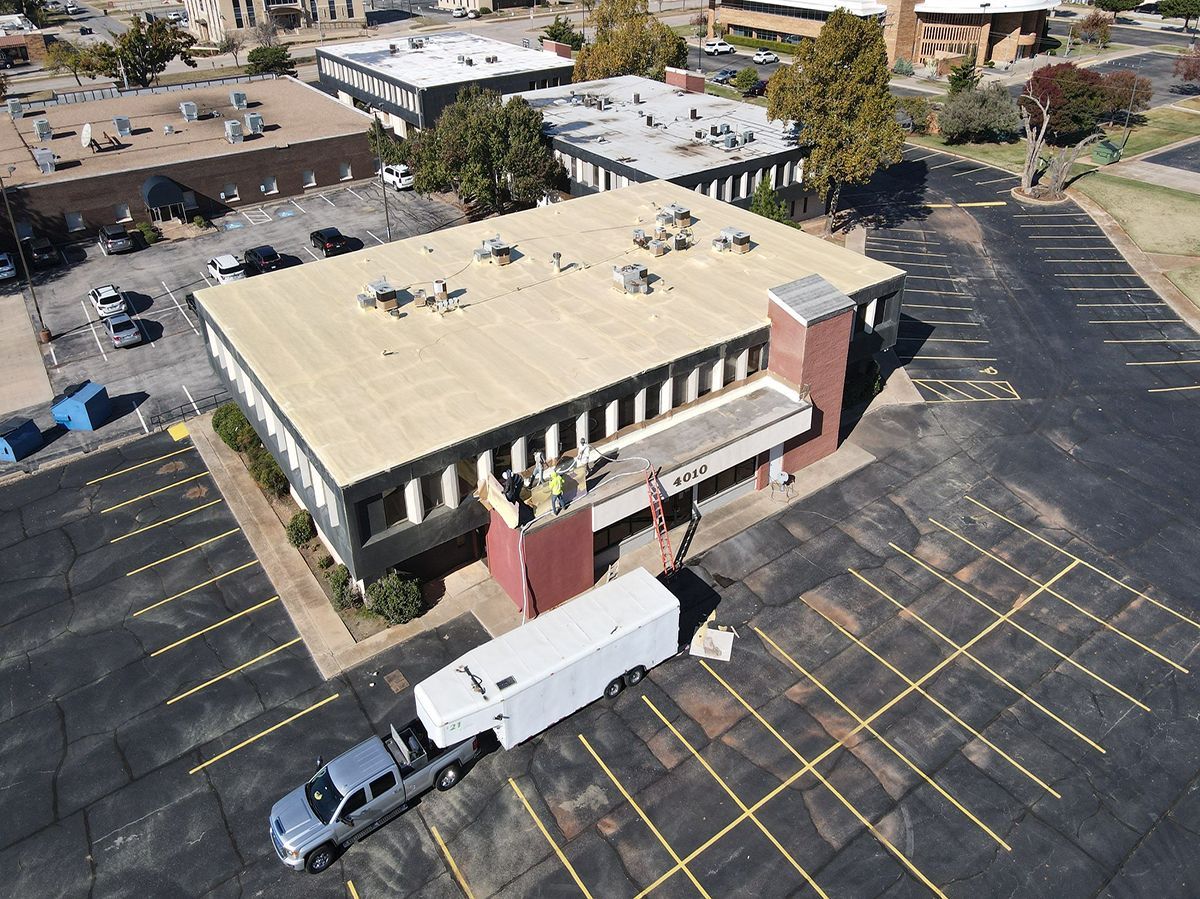 Aerial view: truck towing a white trailer in front of a tan-roofed building with brick accents and a parking lot.