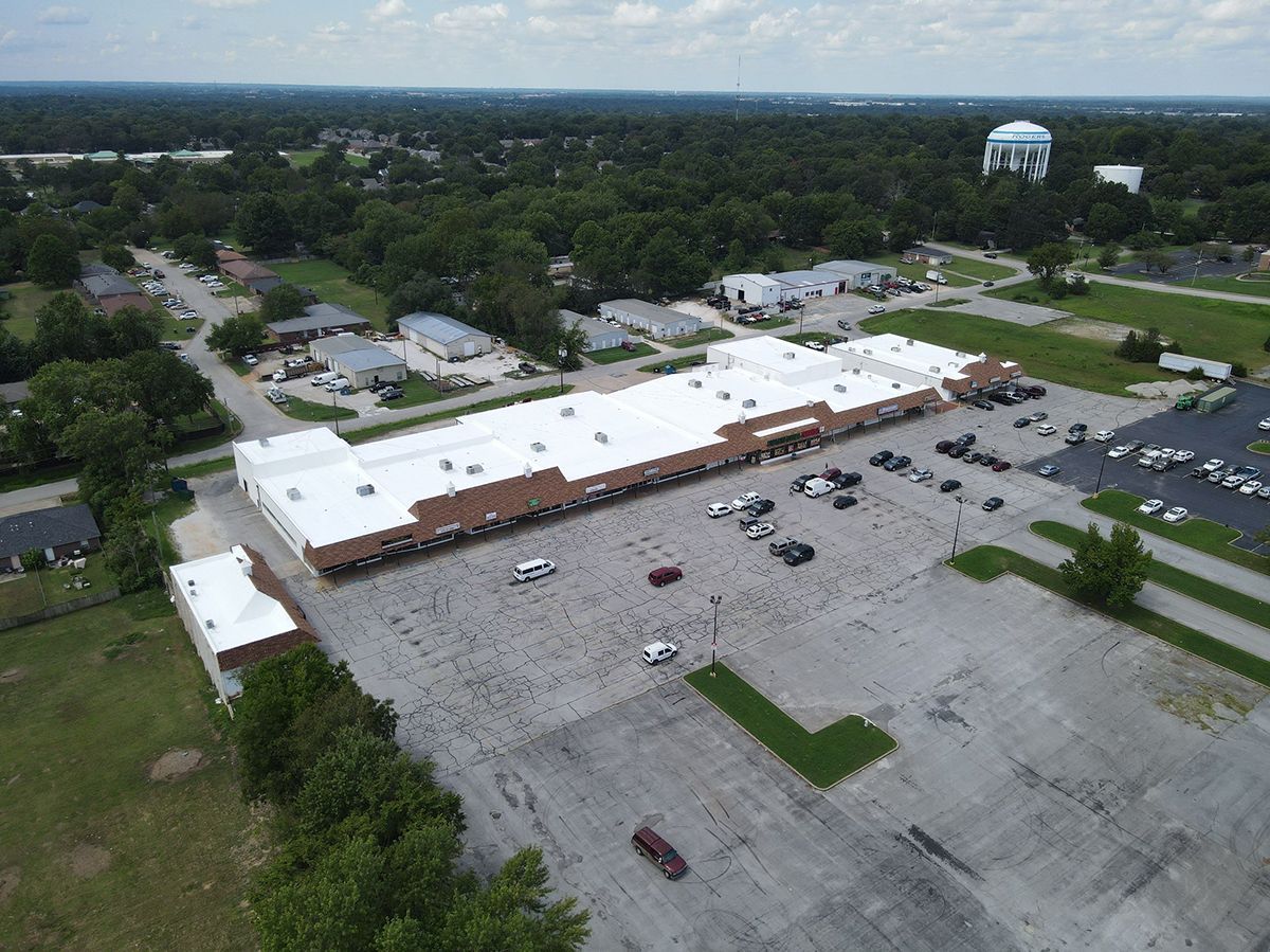 Aerial view of a commercial building with a large parking lot, surrounded by trees and houses, a water tower visible in the distance.