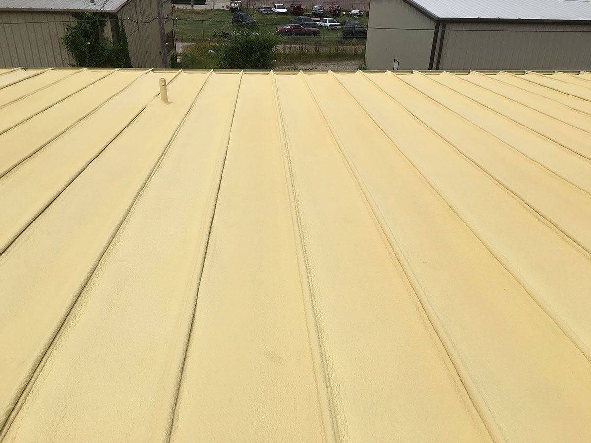 Yellow-coated metal roof with parallel ridges, viewed from above. Buildings and a parking area are in the background.