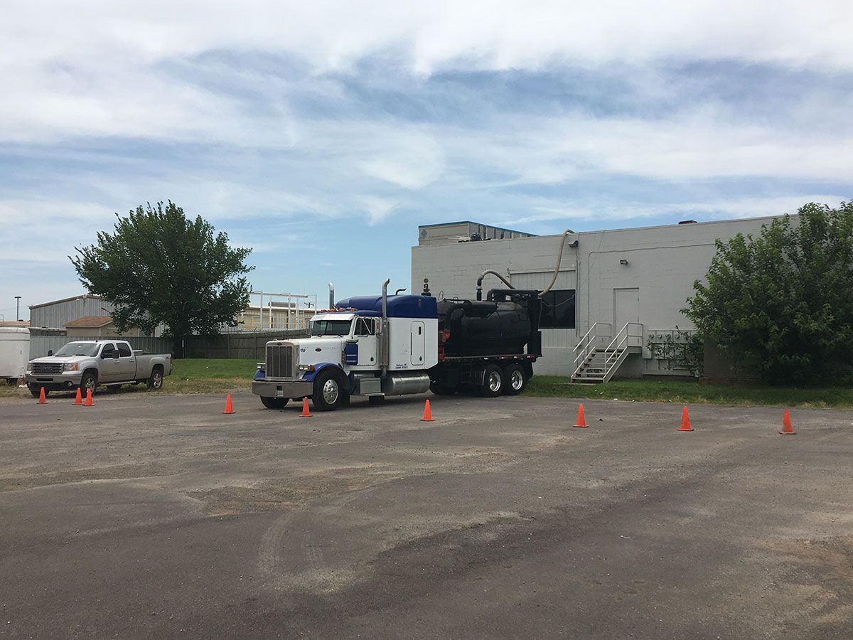 Semi-truck parked near a white building, possibly pumping liquid; orange cones are placed in front of it.