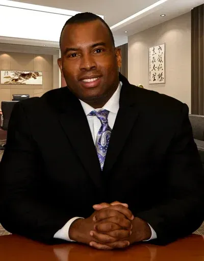 Man in a black suit with a patterned tie, arms crossed, seated at a desk in an office.