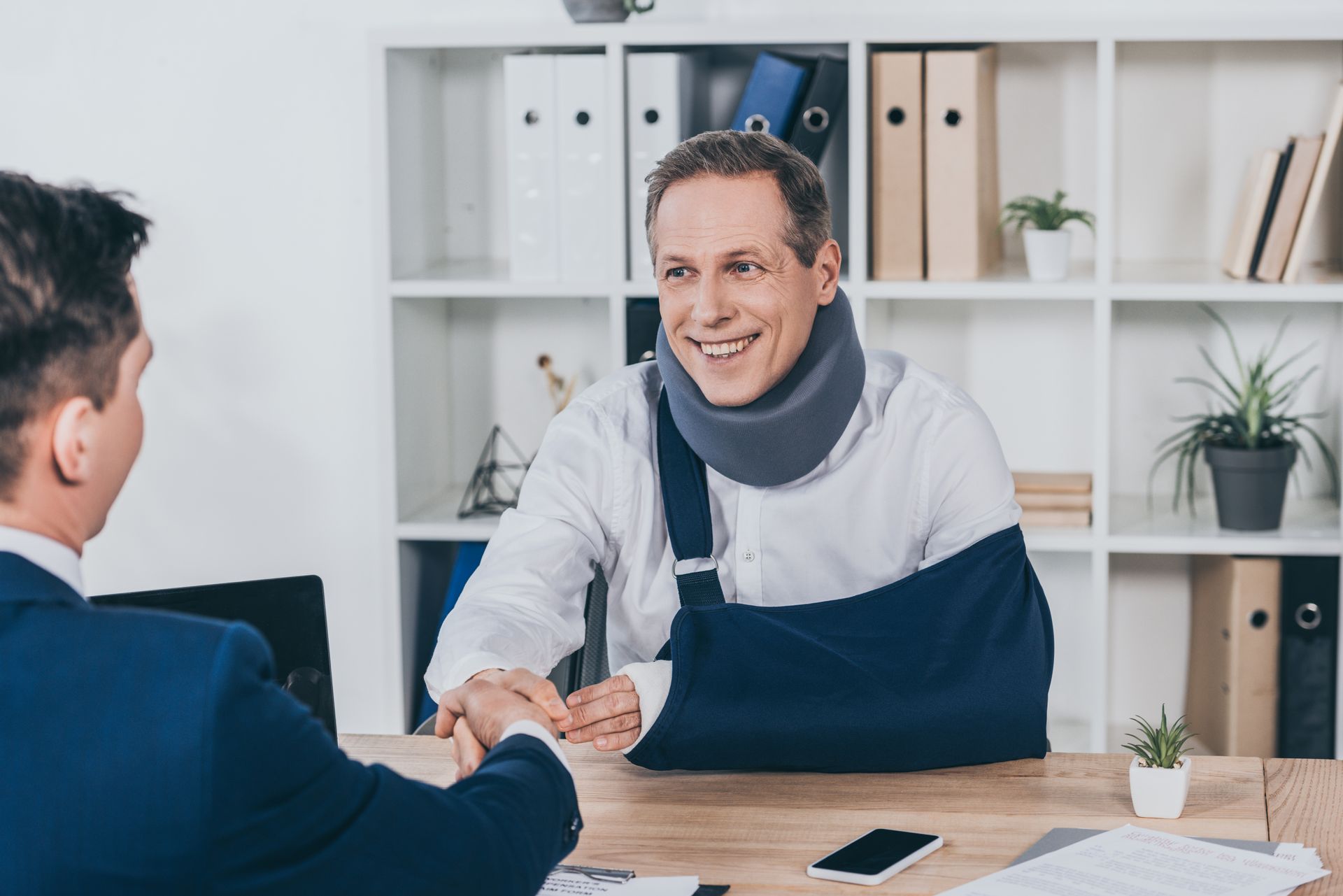 Man in arm sling and neck brace shaking hands with another man at a desk in an office.