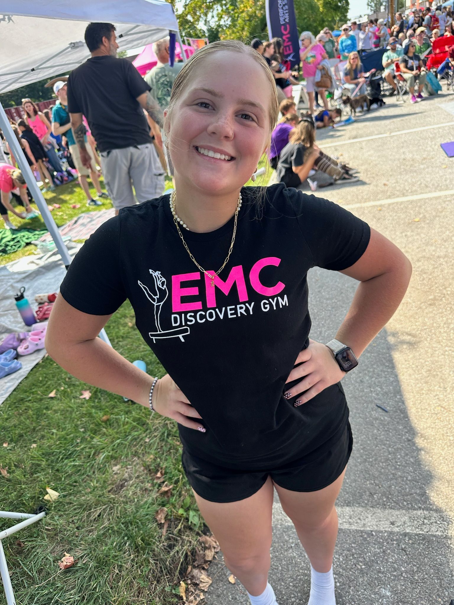 Smiling young woman in black shirt and shorts, hands on hips, at outdoor event.