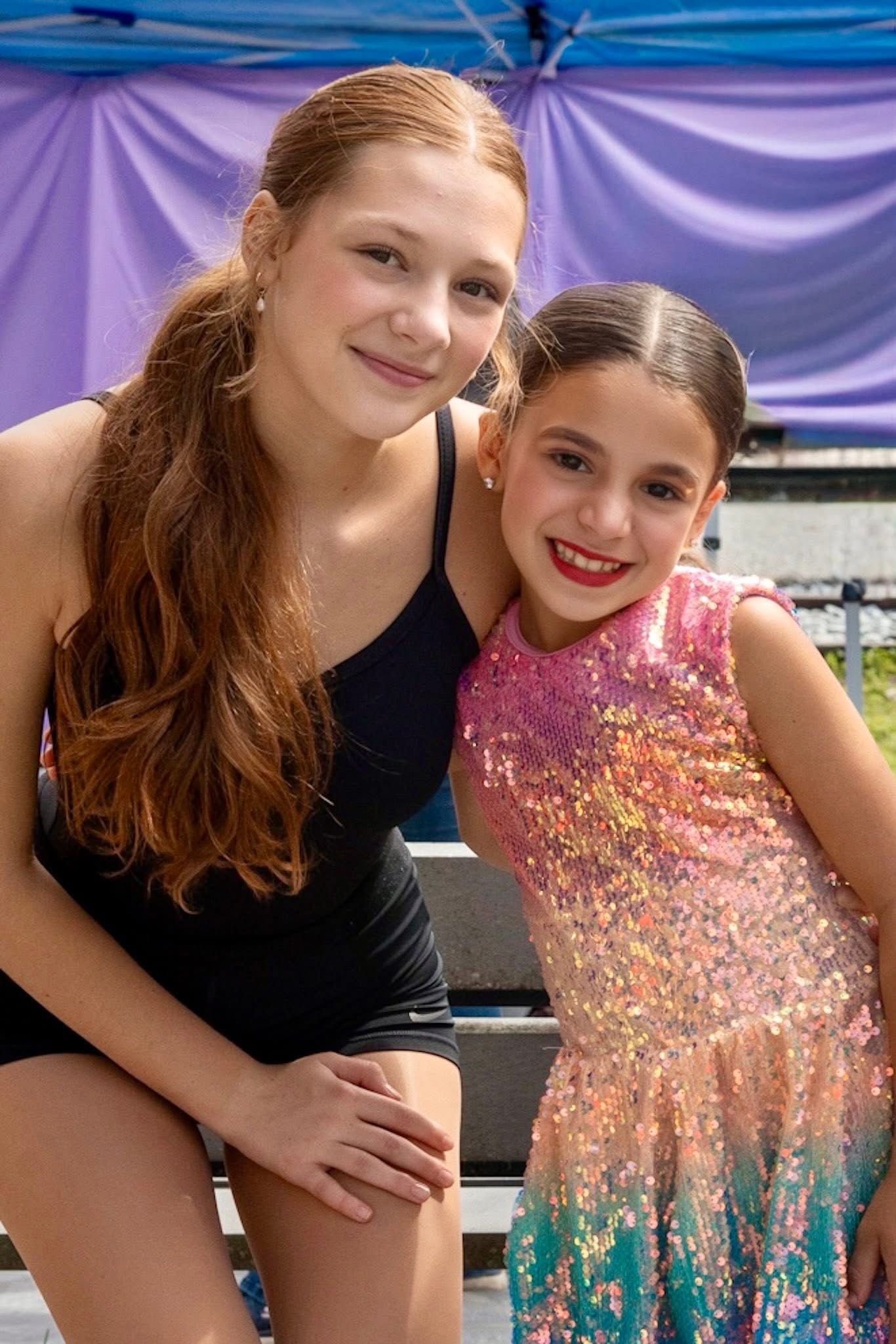 Two young female dancers smiling; one in black, the other in a sparkly costume.
