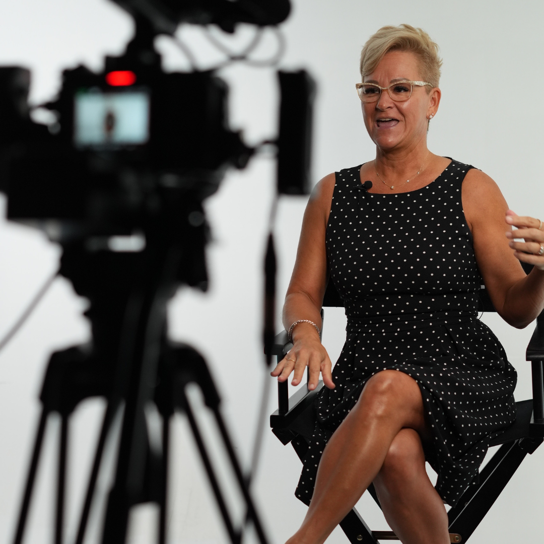 Robin in black dress sitting in a director's chair, arm raised,