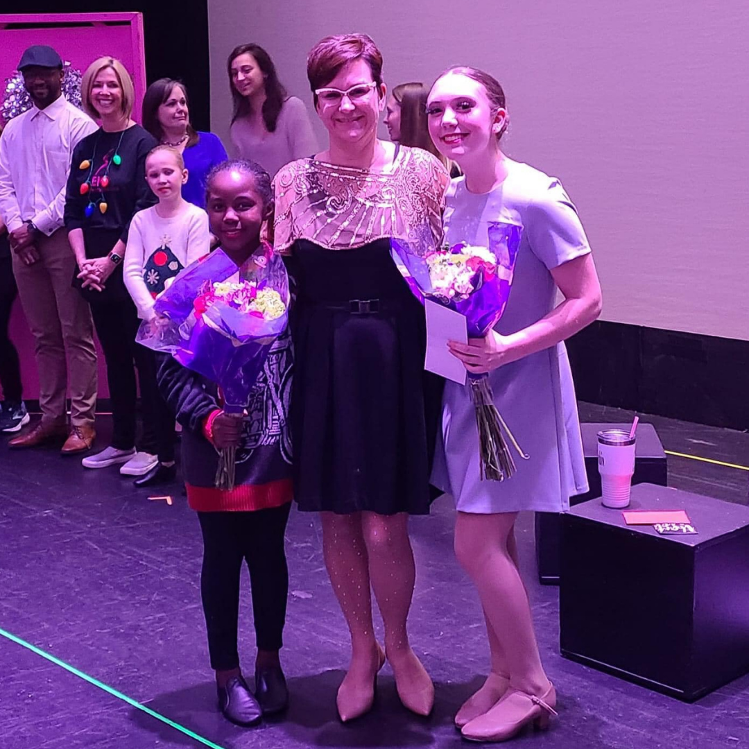 Three people on a stage hold flowers, posing for a photo. Others stand behind them.