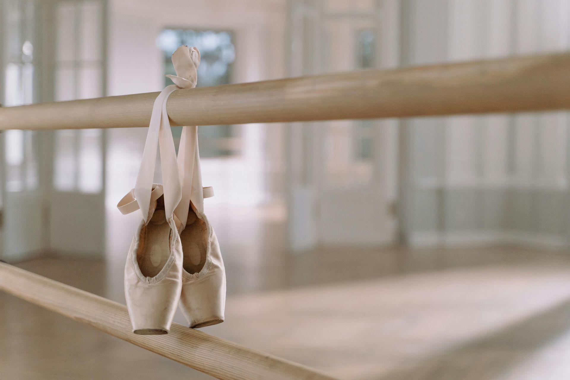 A pair of ballet shoes with ribbons tied, hanging on a wooden barre in a dance studio.