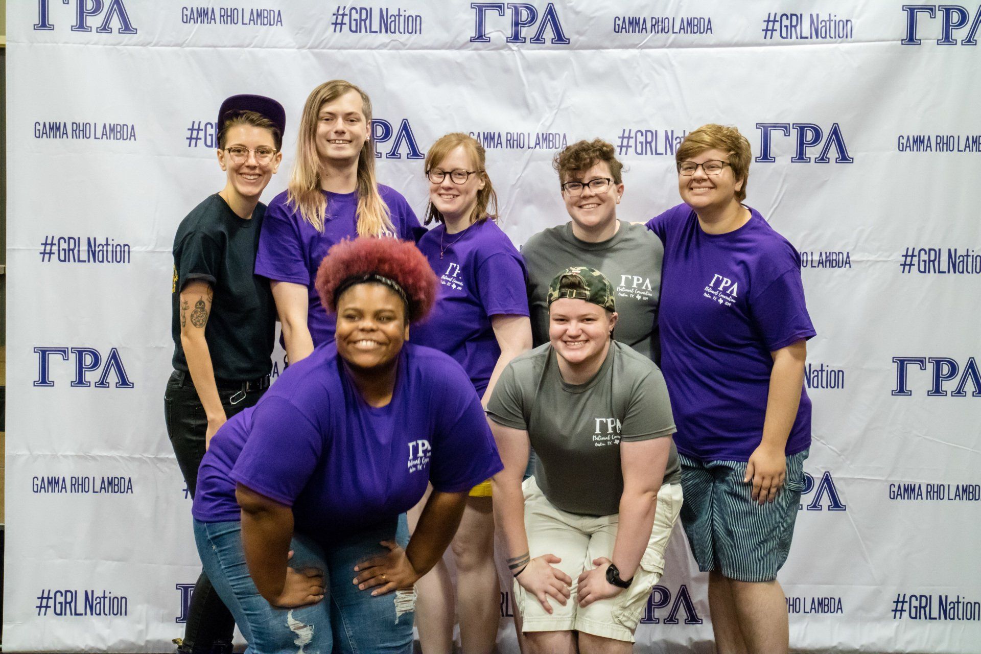 A group of people in purple, gray, and black shirts pose togther in front of a background with text on it.