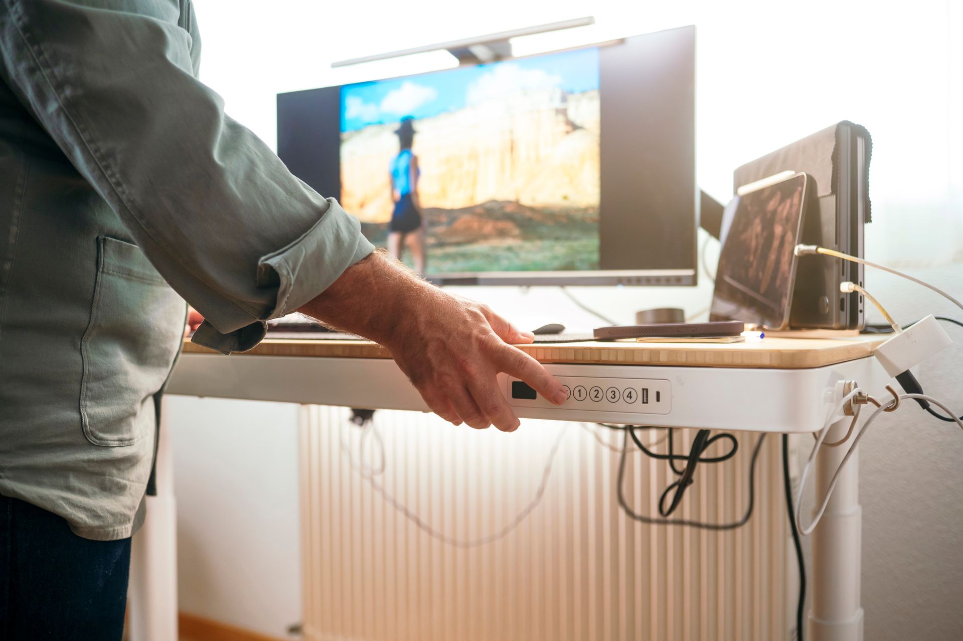 A person adjusts the height of a motorized standing desk in a home office with a computer monitor and laptop.