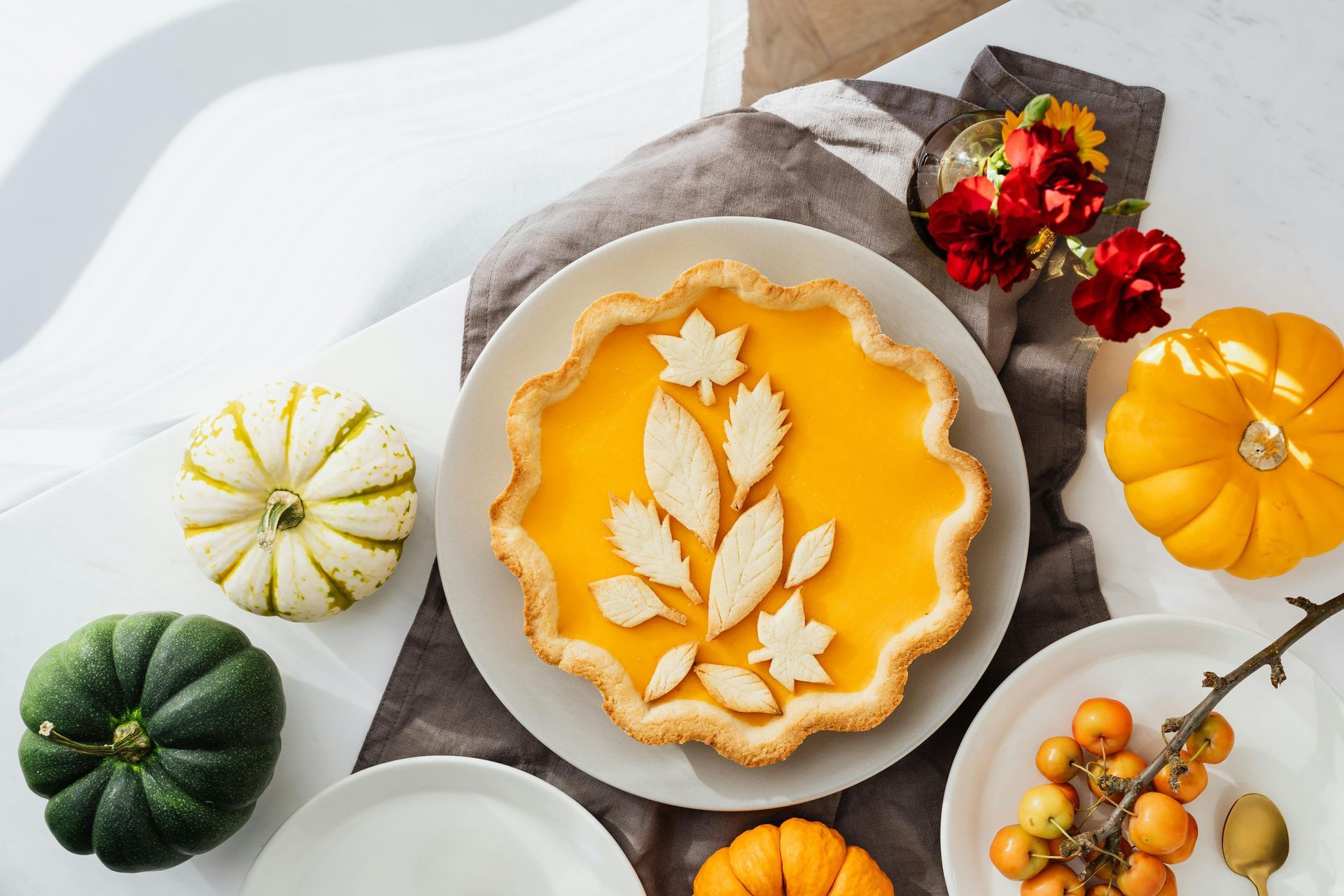Pumpkin pie with leaf decorations, surrounded by gourds, flowers, and plates.