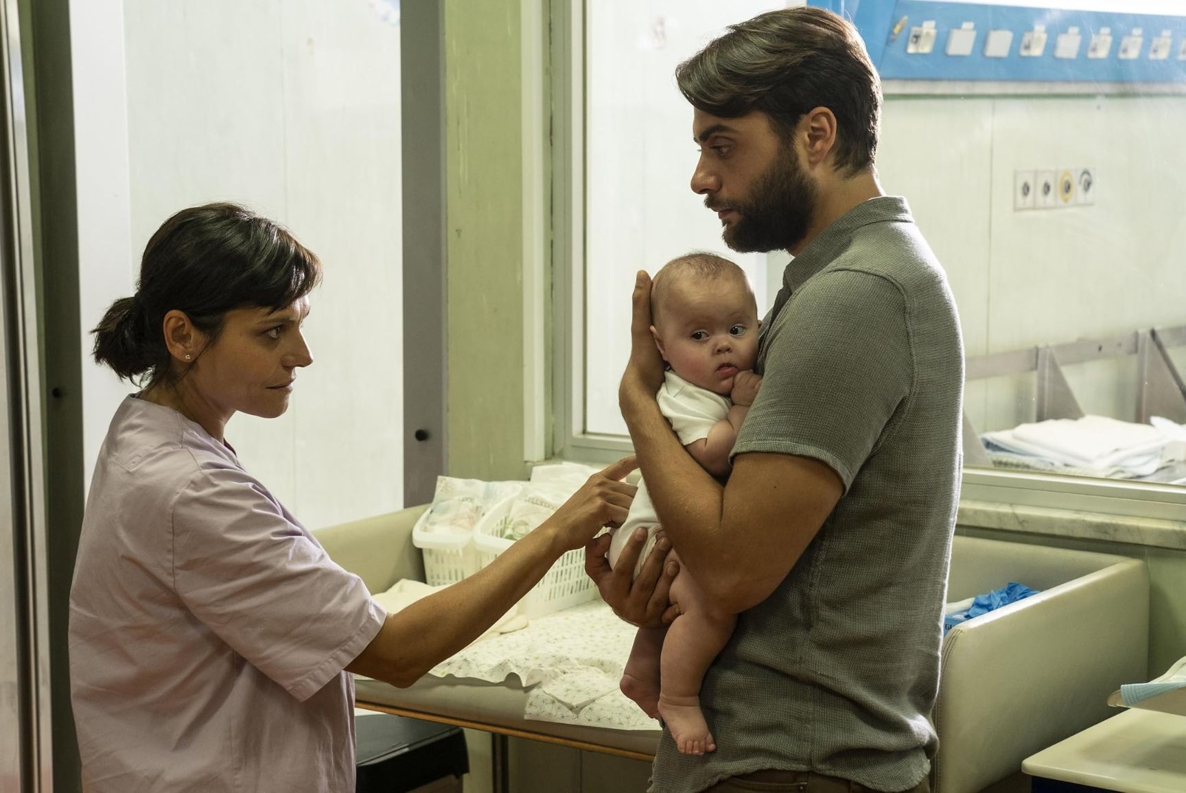 Ein Mann hält ein Baby, während eine Frau in einer rosa Uniform in einem Raum auf das Baby zeigt.