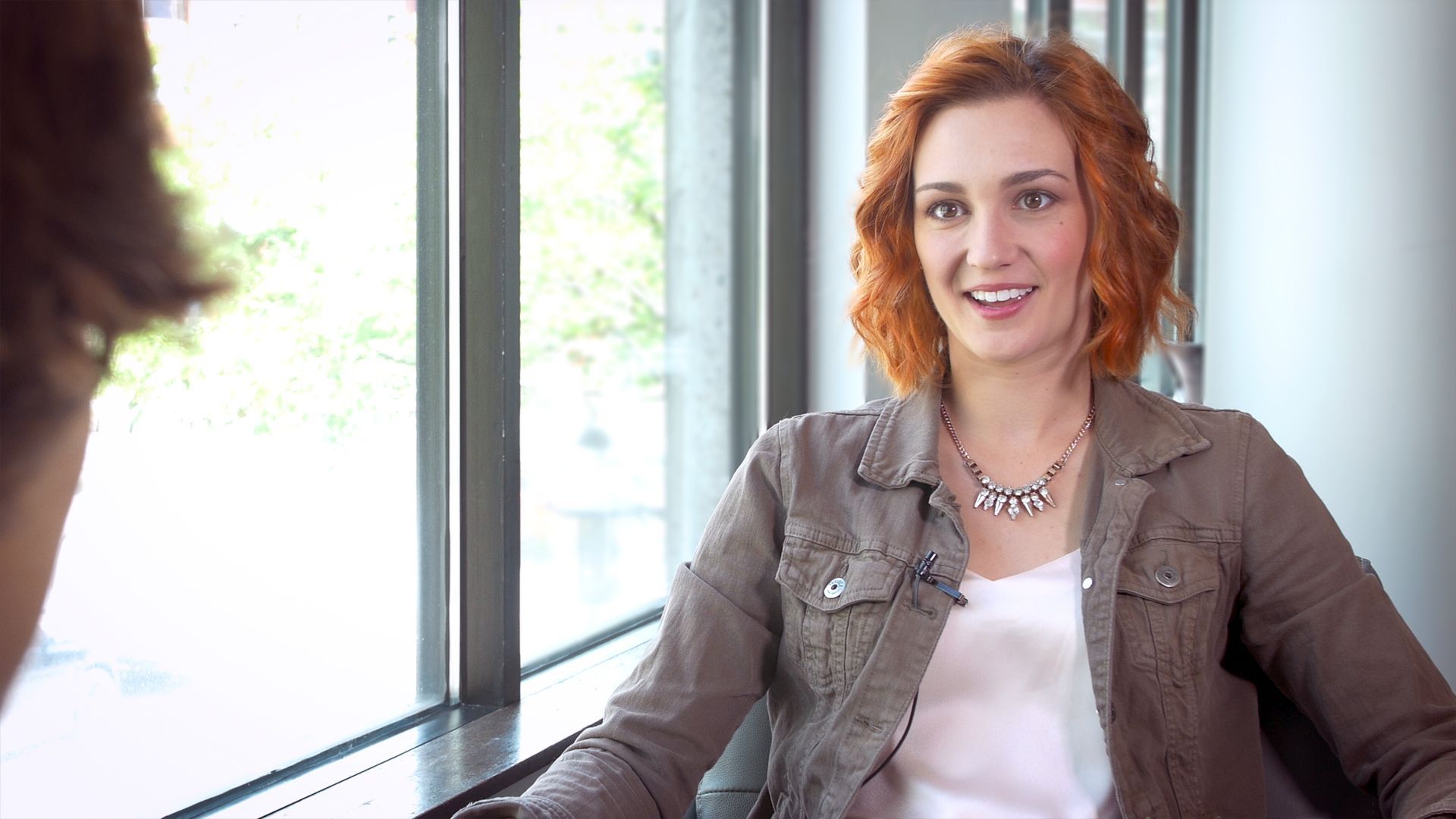 Frau mit roten Locken lächelt während eines Interviews am Fenster; trägt ein weißes Oberteil und eine braune Jacke.