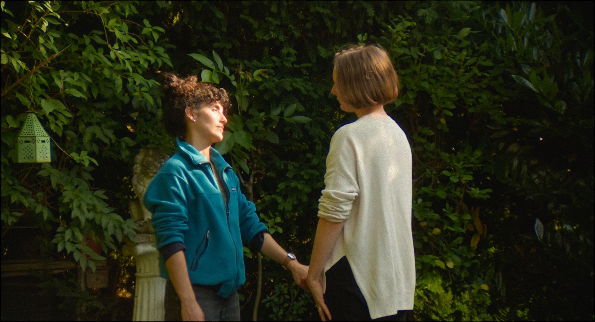 Zwei Frauen halten in einem Garten Händchen; die eine trägt eine blaugrüne Jacke, die andere einen weißen Pullover, beide lächeln.