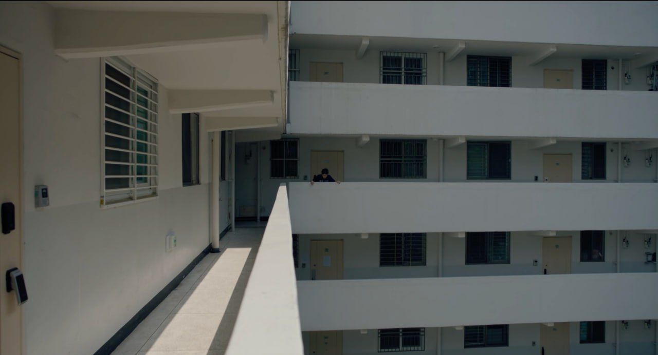 Eine Person steht auf einem weißen Balkon eines mehrstöckigen Gebäudes und schaut hinaus.
