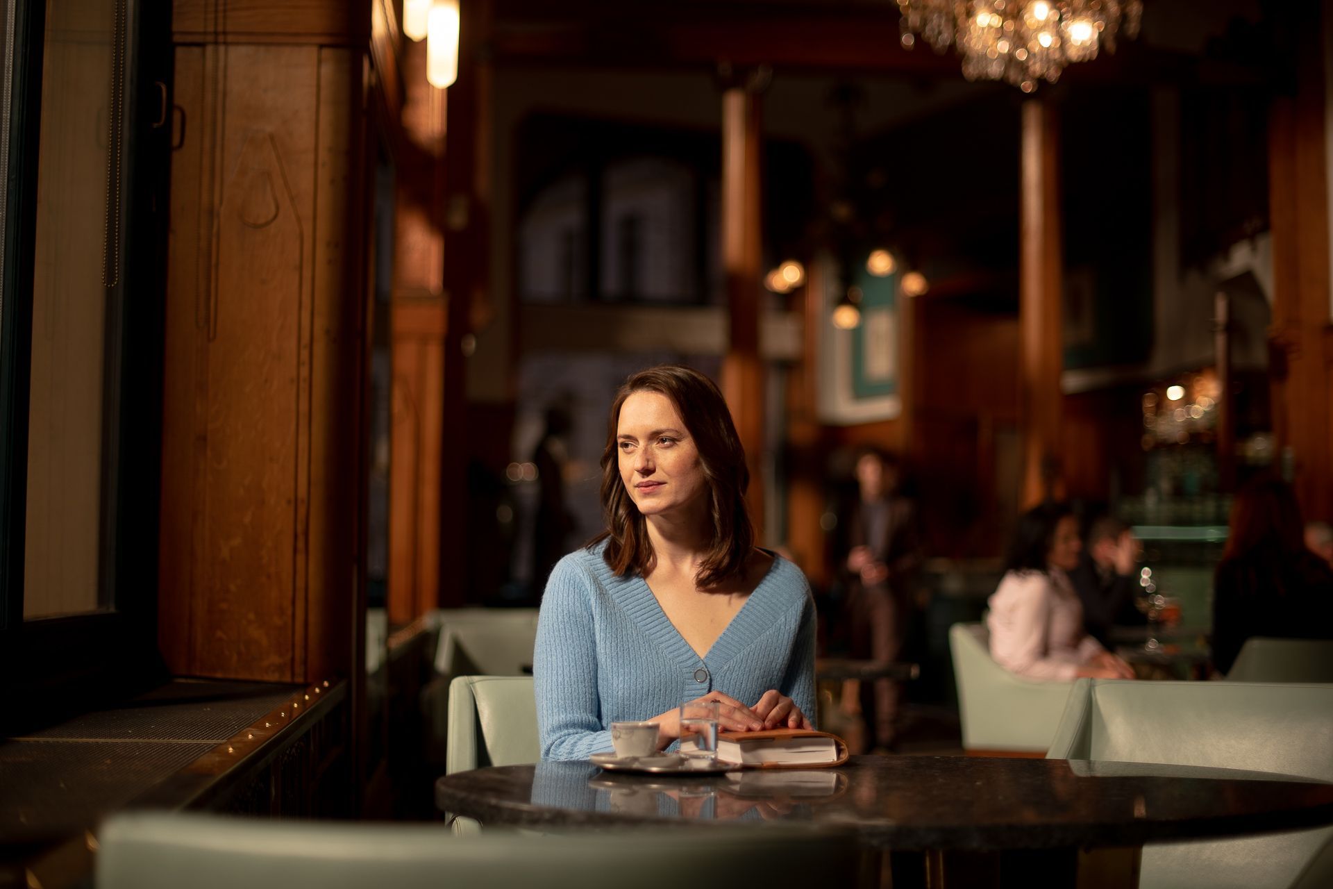 Eine Frau im blauen Pullover sitzt an einem Tisch in einem Café und schaut aus dem Fenster.