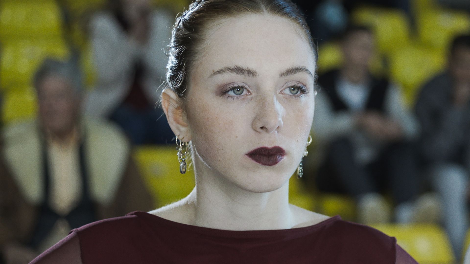 Frau mit dunklem Lippenstift, die aufmerksam blickt. Sie trägt ein dunkelrotes Kleid und Ohrringe, vor einer verschwommenen Menschenmenge.