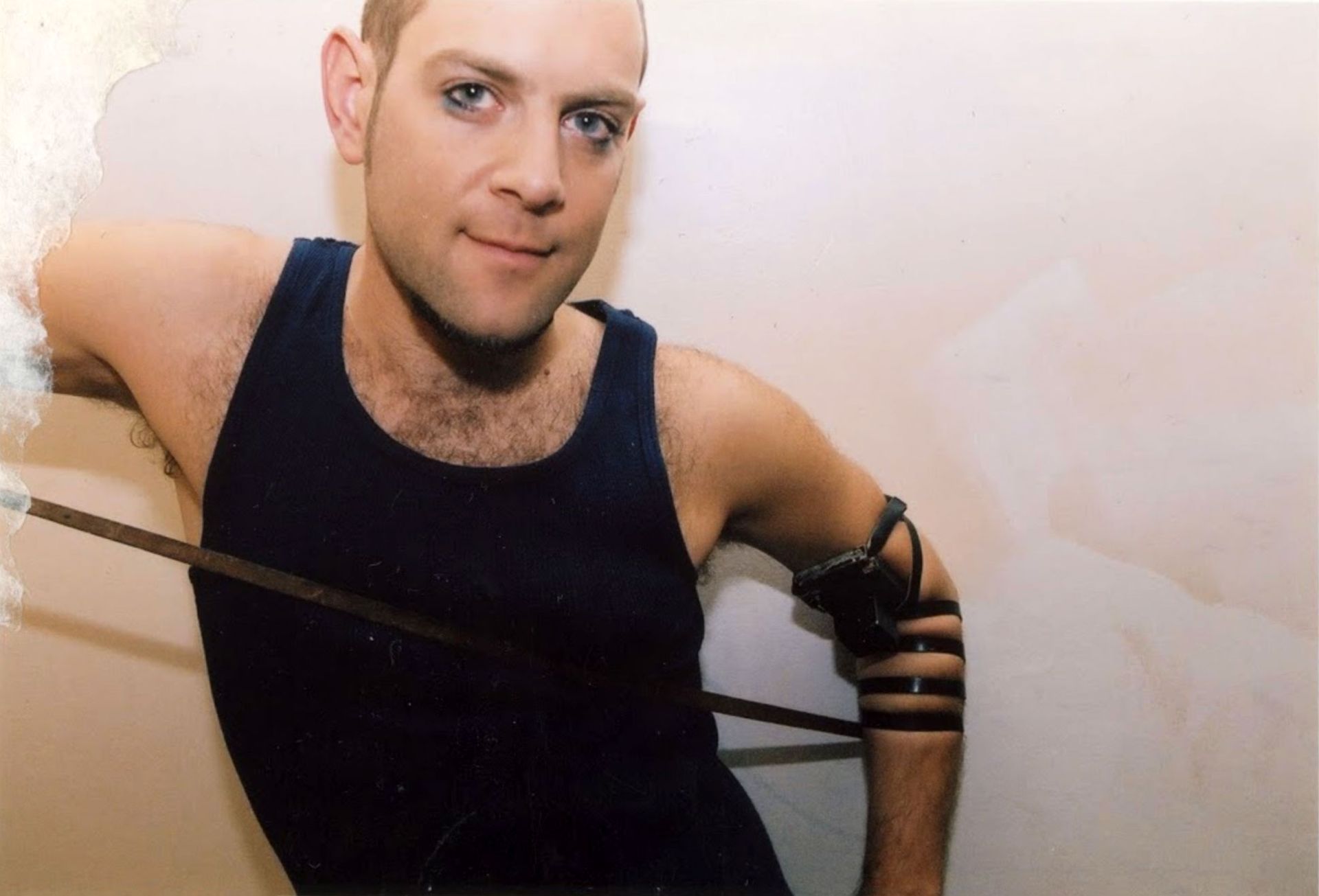 Mann im dunklen Tanktop lehnt mit schwarzen Armbinden an der Wand.