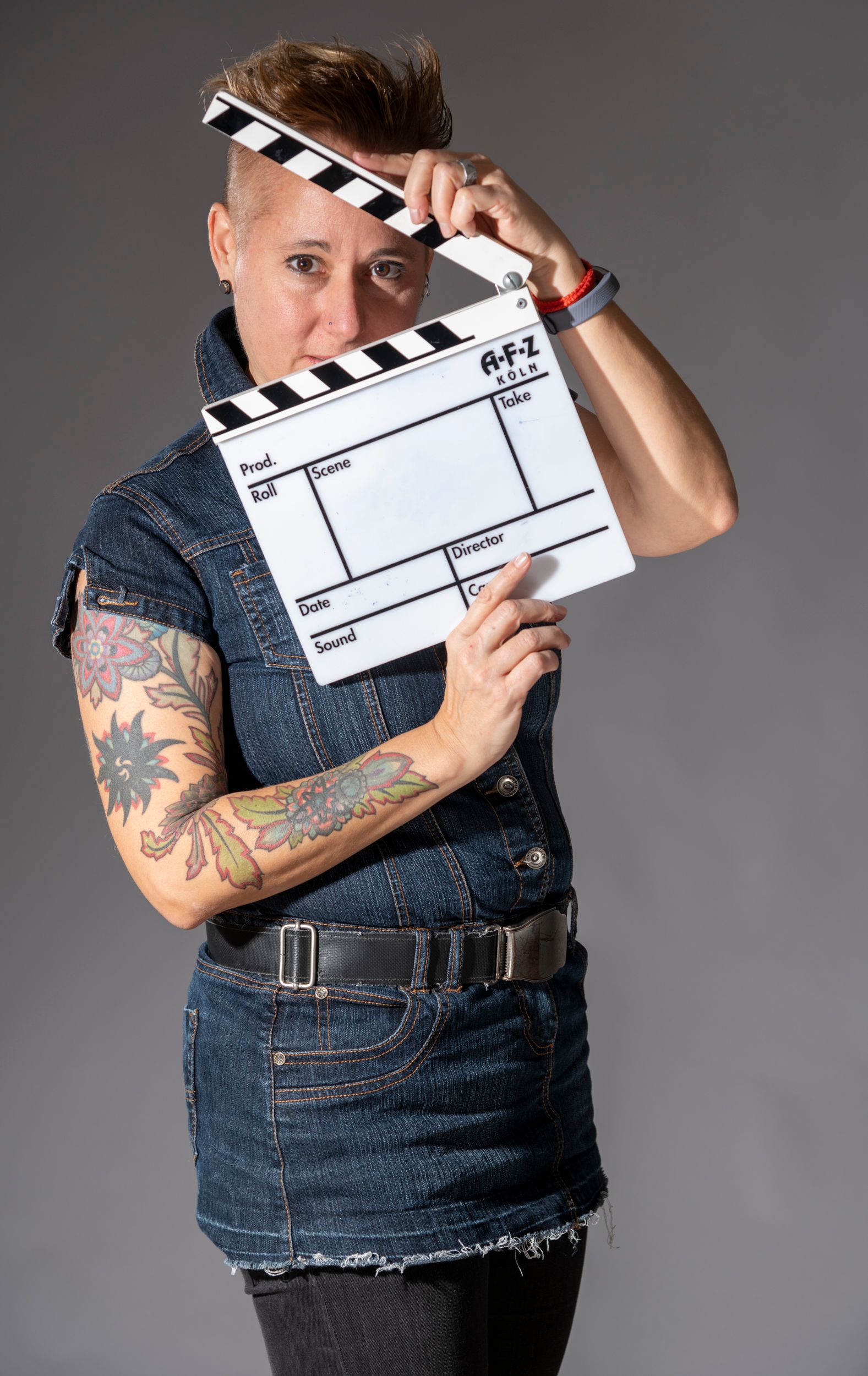Eine Frau mit Tattoos hält eine Filmklappe vor ihr Gesicht