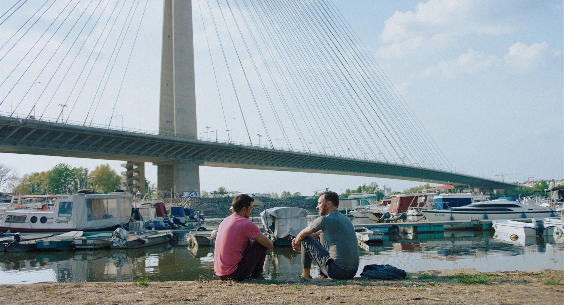 Zwei Männer sitzen am Ufer eines Gewässers vor einer Brücke