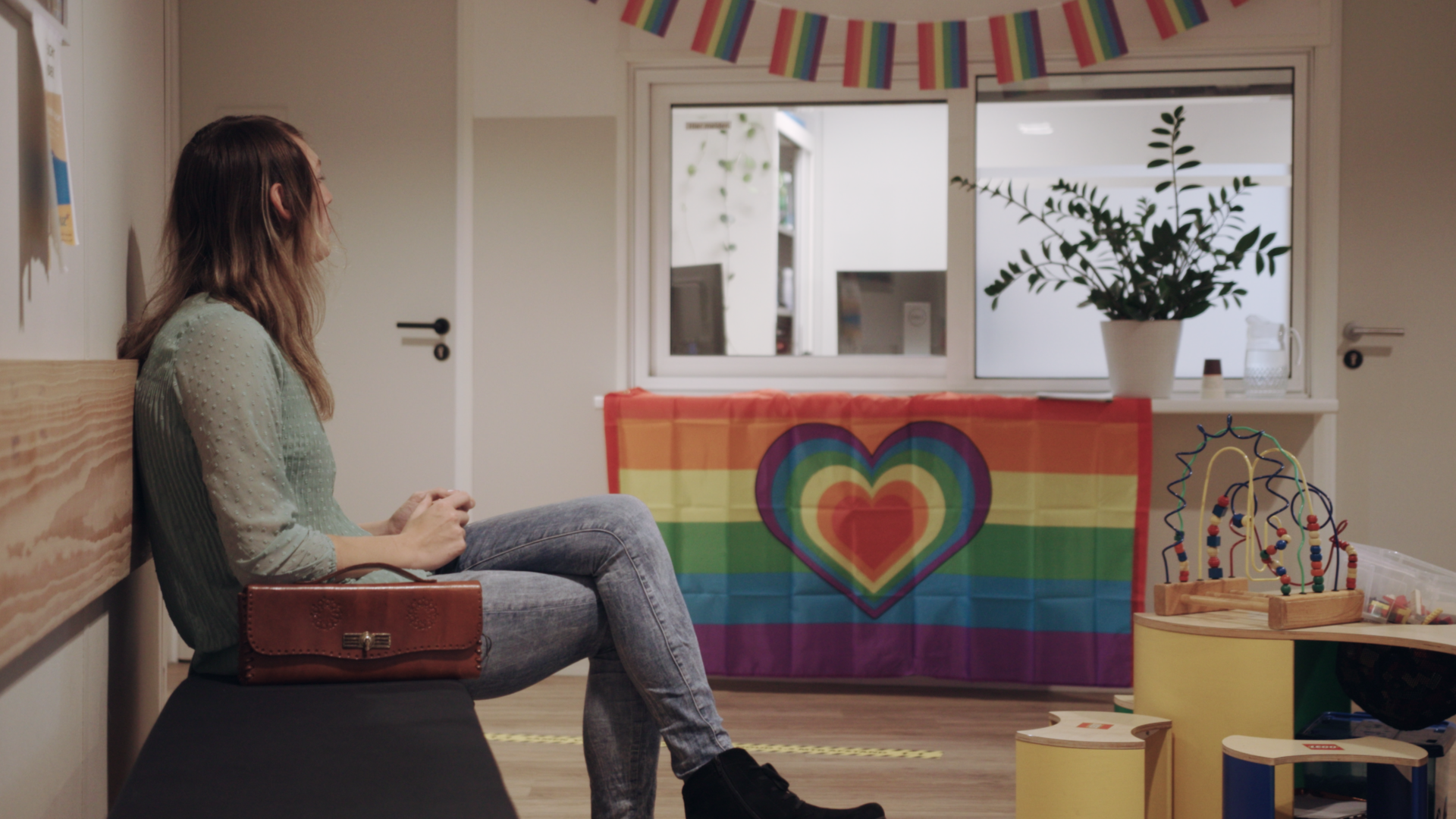 Eine Person sitzt nachdenklich in einem Wartezimmer mit LGBTIQ+-Dekor.