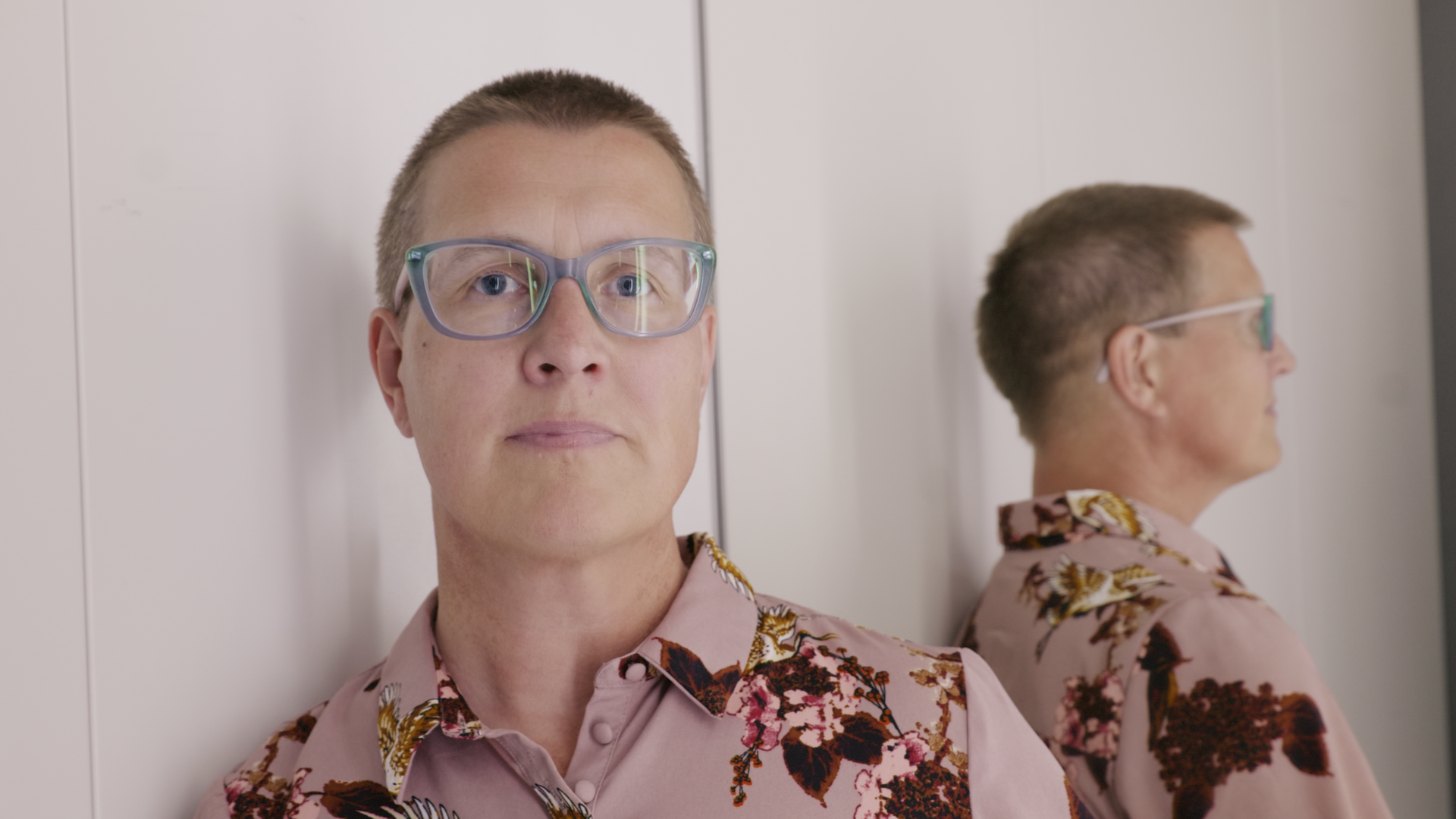 Eine Person mit Brille und rosa geblümtem Hemd lehnt an einer weißen Wand und wird in einem Spiegel reflektiert.
