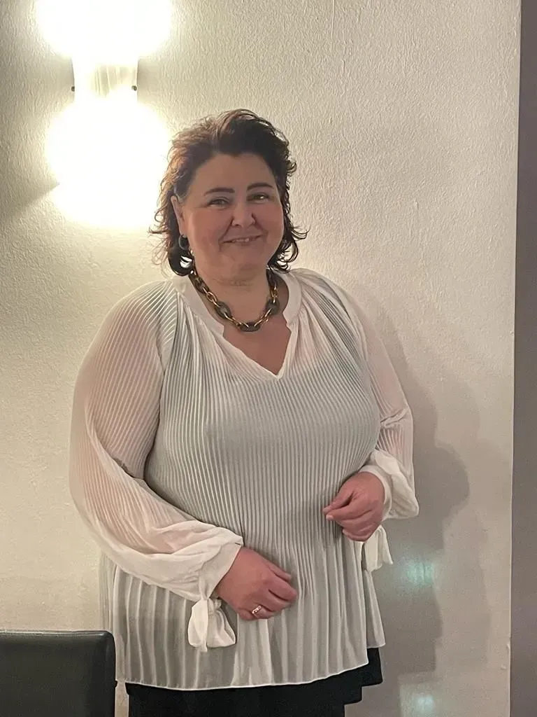 Eine Frau steht vor einer Wand und trägt ein weißes Oberteil und eine Halskette.