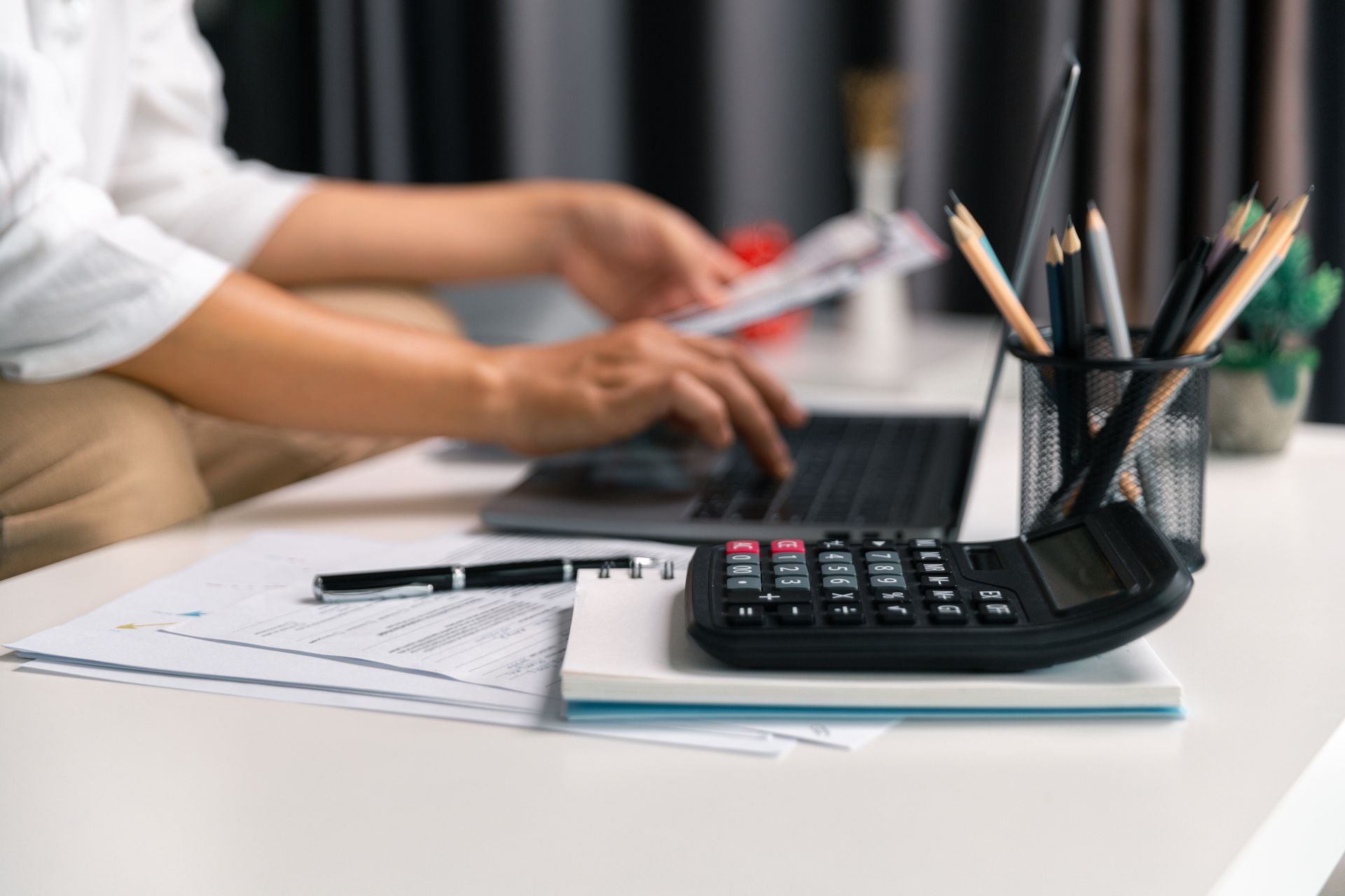 Person typing on laptop, reviewing papers with calculator, pencils, and notepad on a table.