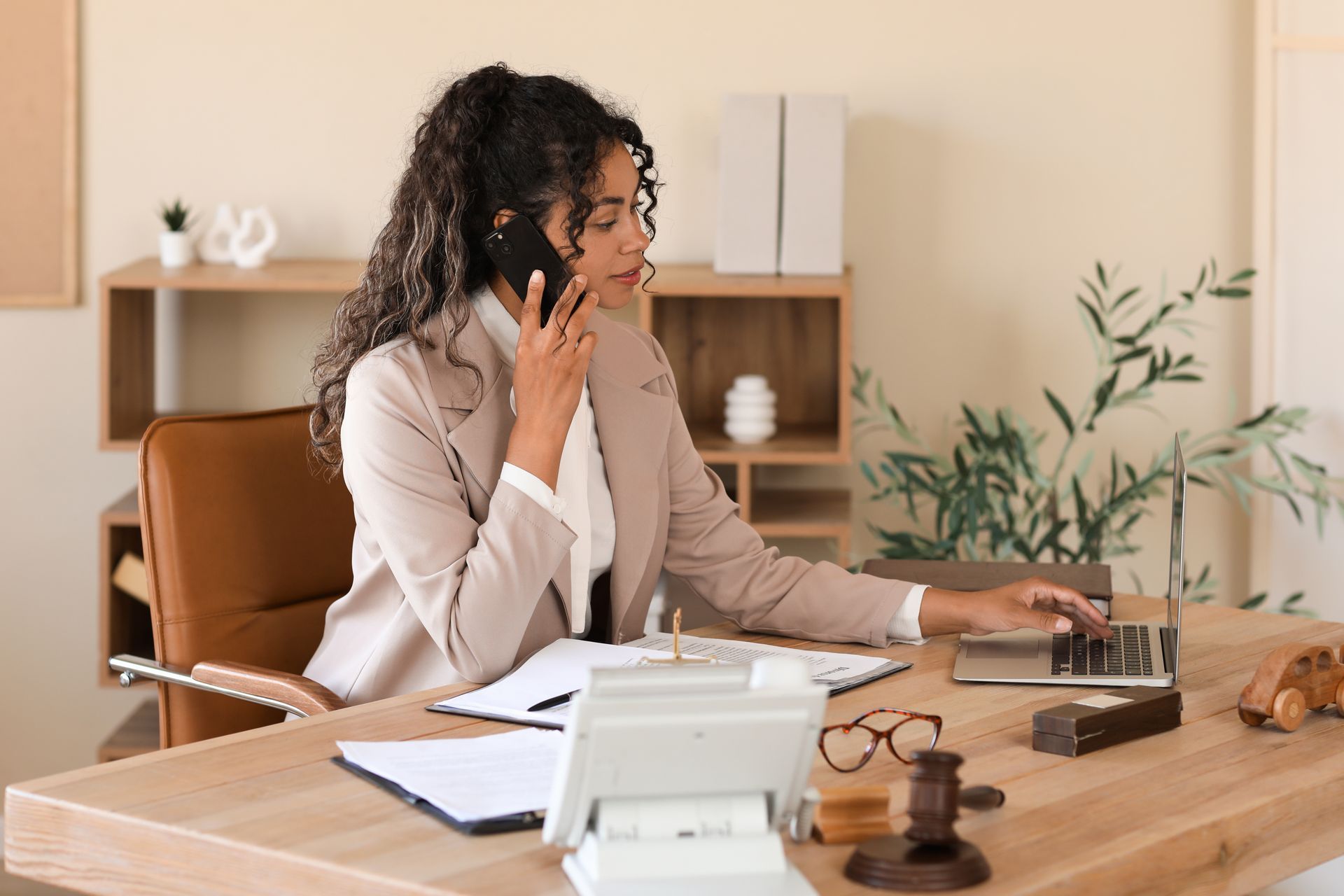 Woman in beige blazer on phone, using laptop at desk, documents, and other office supplies.