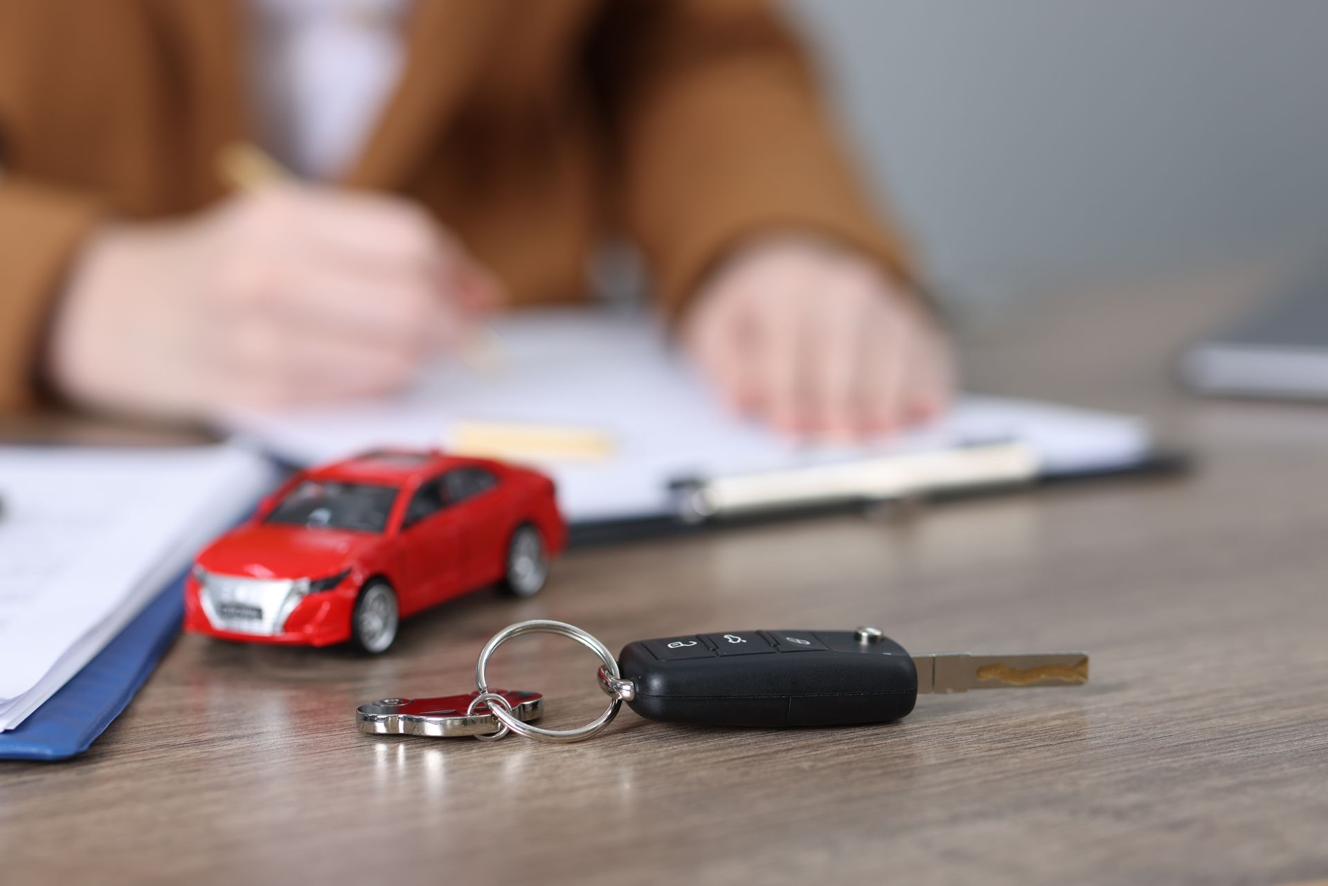 Car keys, toy red car, and paperwork on a desk; a person is signing documents in the background.