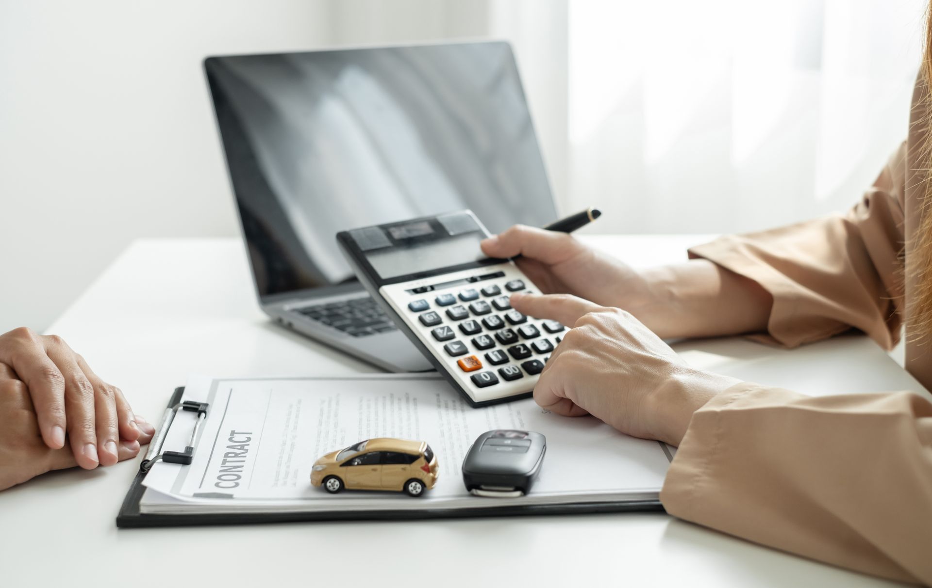 Person calculating car loan details with a calculator, a laptop, and a contract in a light-filled office.