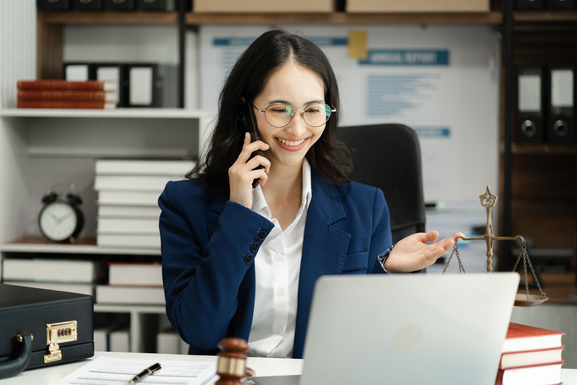 Woman in suit on phone, smiling at laptop in an office setting. Scales of justice on desk.