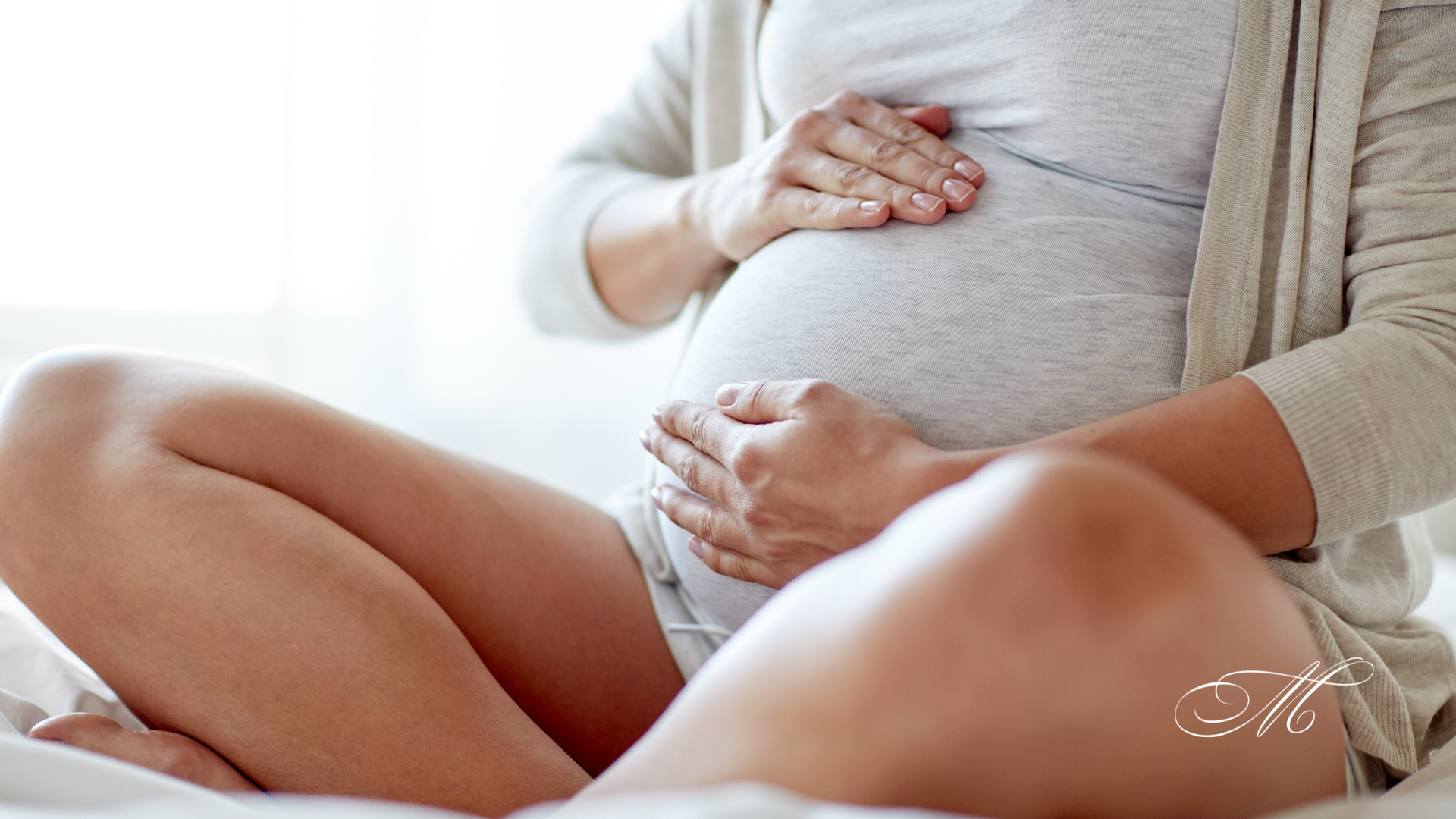 A pregnant woman is sitting on a bed holding her belly.