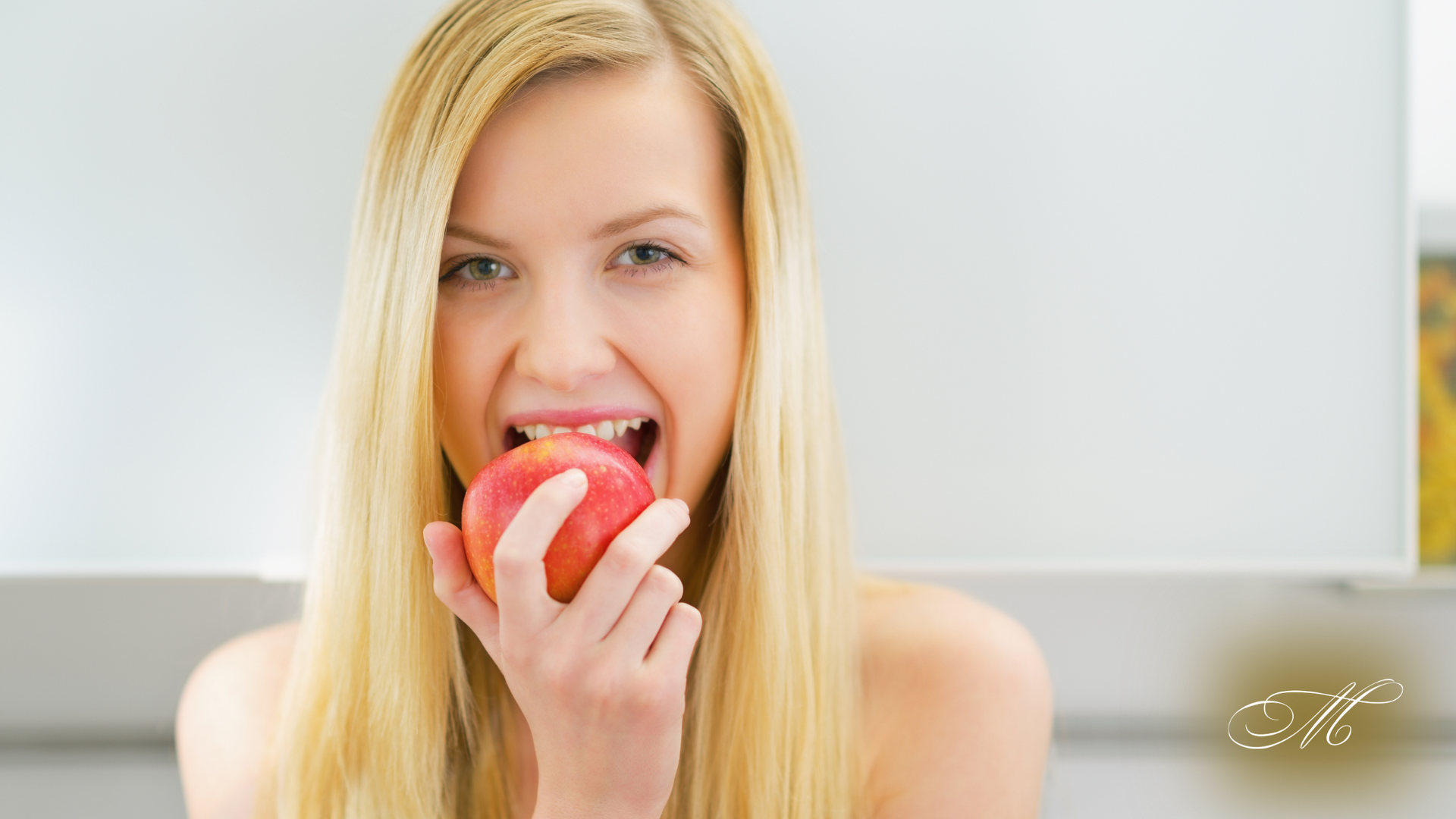 Blonde woman taking a bite of a red apple indoors, looking directly at the viewer.