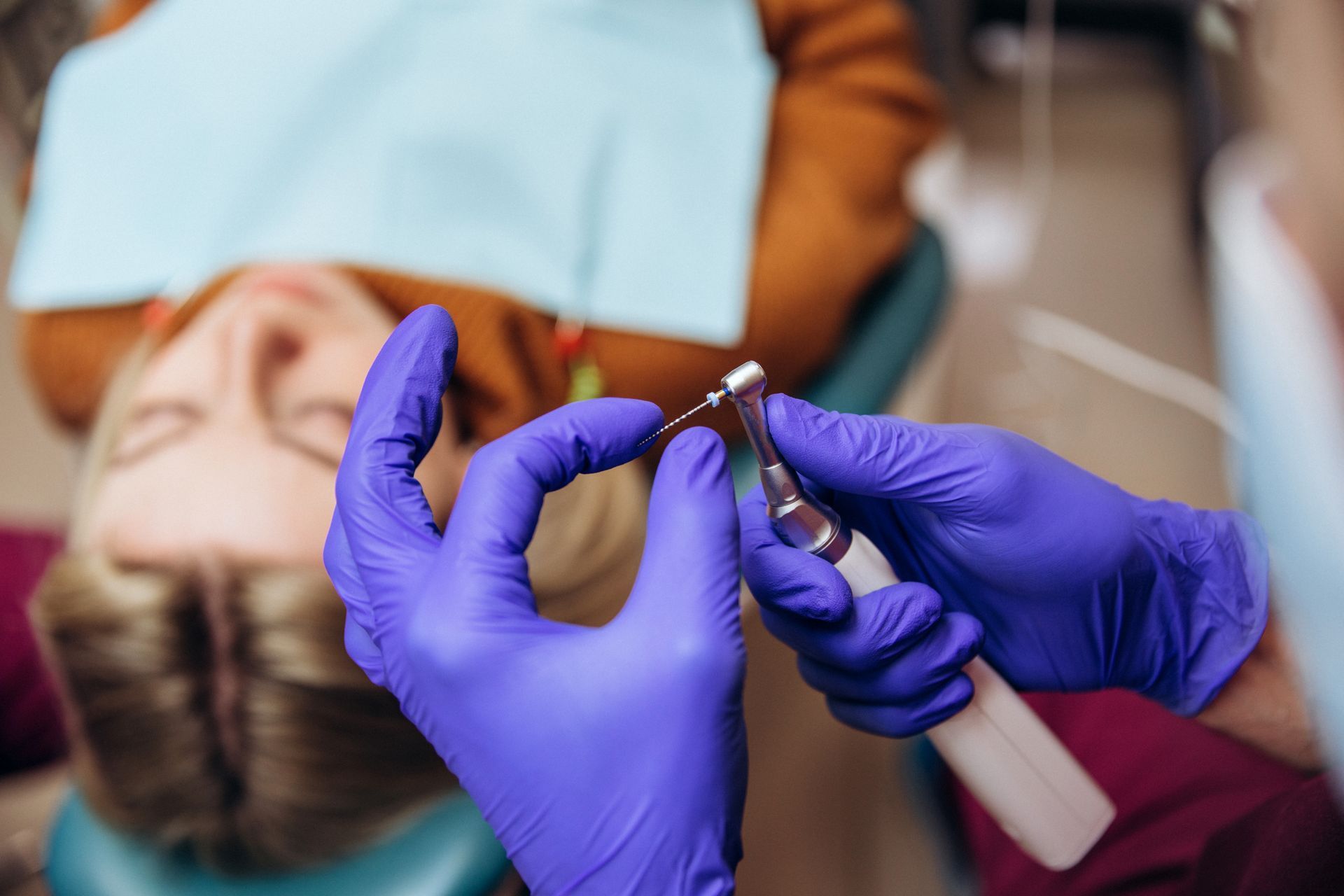 A dentist wearing blue gloves holds a small endodontic file attached to a handpiece near a patient in a dental chair.
