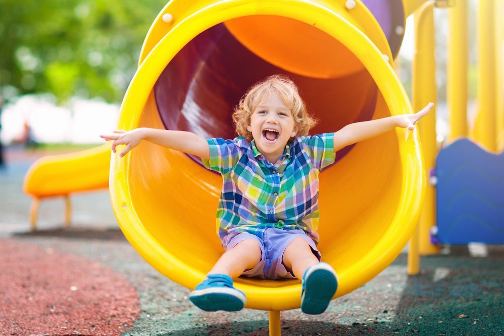 A little boy is sitting in a yellow slide at a playground.