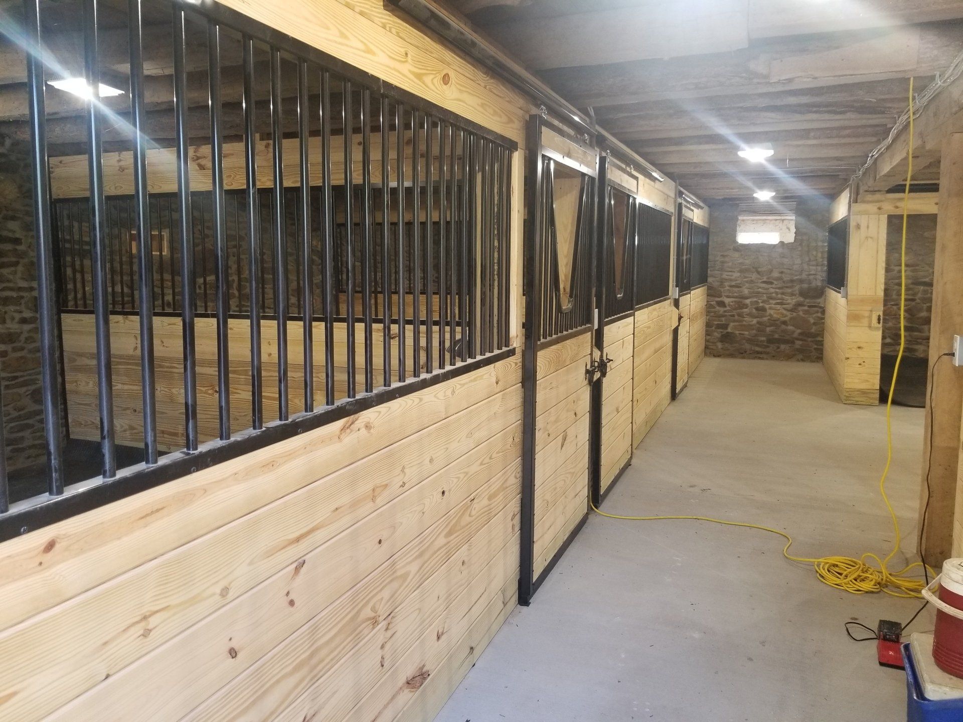 Horse stalls lined along a corridor in a stable, with wooden walls, black metal bars, and a concrete floor.