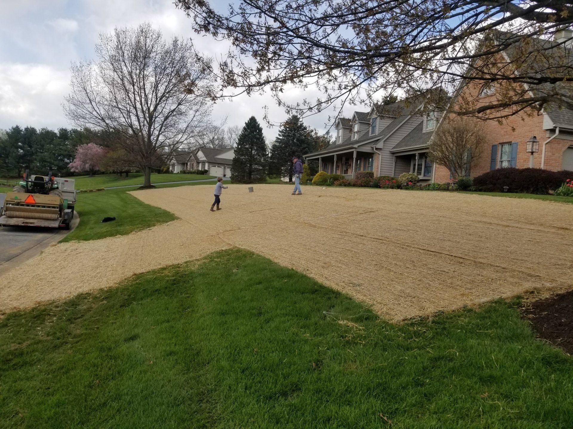 Gravel driveway with two people playing. Green grass borders the gravel. A residential neighborhood on a cloudy day.